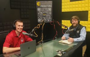 Two hosts with headphones sit at a podcasting table with microphones