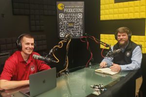 Two hosts with headphones sit at a podcasting table with microphones