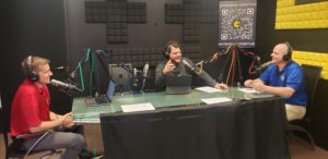 Three individuals sit at a podcasting table with microphones